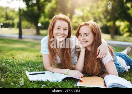 She is my partner in crime. Joyful attractive redhead girls with cute freckles hugging while lying on grass in warm summer day, doing homework and Stock Photo