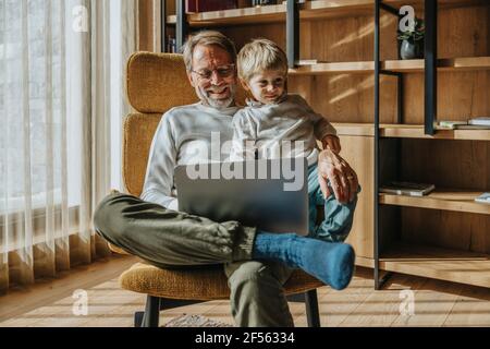 Smiling mature man with son using laptop while sitting on chair Stock Photo