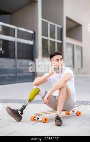 Young disabled man talking through mobile phone while sitting on skateboard Stock Photo