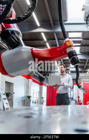 Robotic welding torch while young man working with laptop in factory Stock Photo