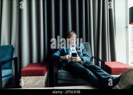 Serious businessman using mobile phone while resting on armchair in cafeteria at office Stock Photo