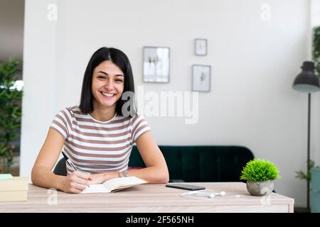 Cheerful woman with notebook and pen sitting at table in living room Stock Photo