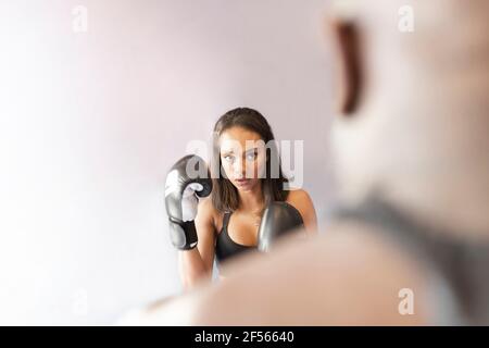 Young woman wearing boxing gloves looking at fitness instructor in gym Stock Photo