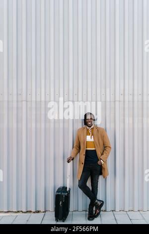 African man with suitcase standing while leaning against gray wall Stock Photo