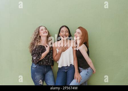 Carefree young women laughing while standing against green wall Stock Photo