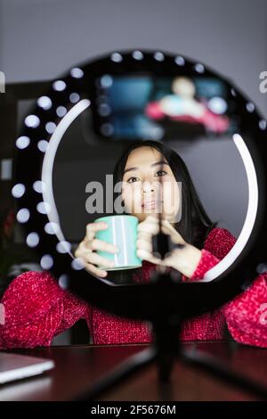 Female influencer holding coffee cup while vlogging in front of ring light Stock Photo