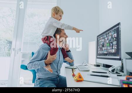 Mature businessman attending video conference while son sitting on his shoulder pointing on computer screen at home office Stock Photo