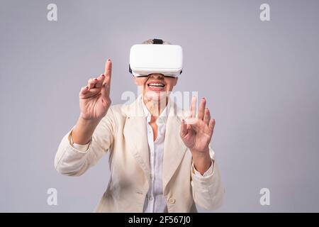 Senior businesswoman gesturing while using virtual reality headset while standing against gray background Stock Photo