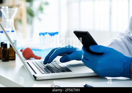 Scientist holding smart phone while using laptop at desk in laboratory Stock Photo