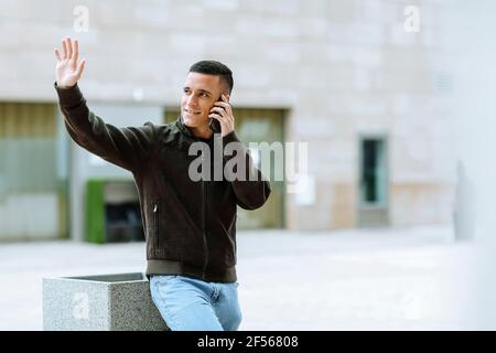 Smiling man waving while talking on mobile phone against building Stock Photo