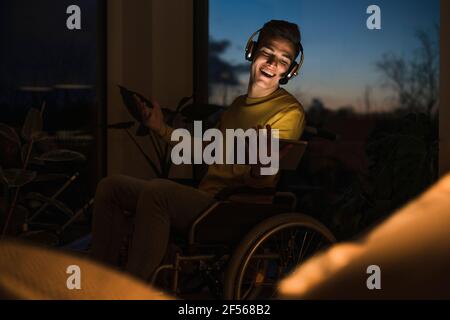 Happy ýoung man in wheelchair wearing wireless headphones while making video call through digital tablet in living room Stock Photo