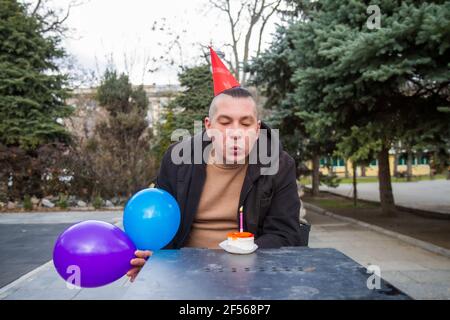 A sad birthday boy wearing a homemade party cap with balloons and cake with a candle celebrates his birthday alone in an empty park during an epidemic Stock Photo