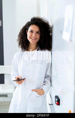 Smiling female scientist with smart phone leaning by whiteboard at laboratory Stock Photo