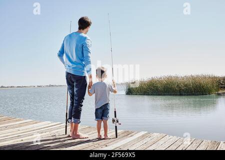 Father and son holding hands while standing with fishing rods on jetty Stock Photo