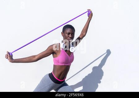 Young female athlete stretching arms with rubber band against white wall during sunny day Stock Photo