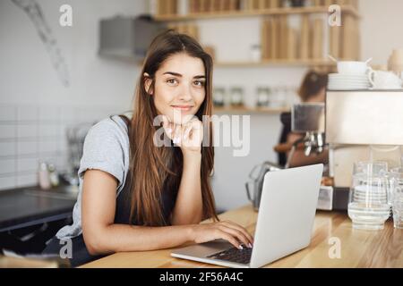 Female cafe owner using laptop computer waiting for first client in the morning looking at camera smiling. Stock Photo