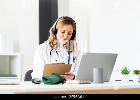 Female medical worker writing on clipboard while giving online consultation through laptop in medical clinic Stock Photo