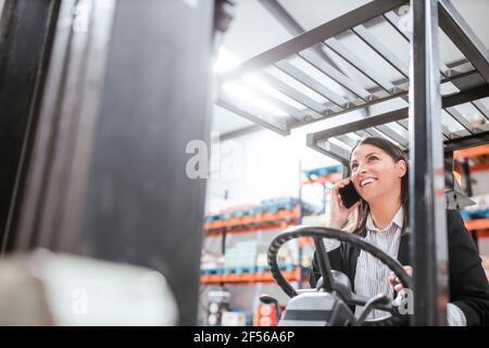 Cheerful female manager talking on mobile phone while operating forklift at warehouse Stock Photo