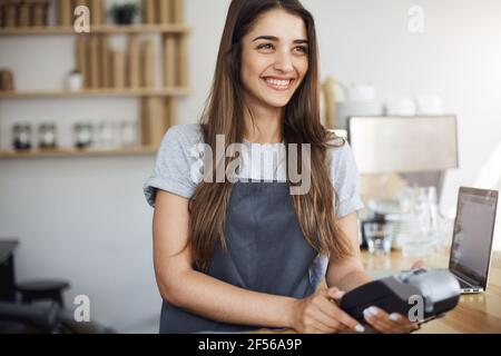 Young female coffee shop employee using a credit card reader and a laptop to bill the customer Stock Photo