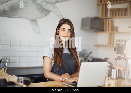 Young female barista using a laptop computer at her work in cafe. Happy employee looking at camera smiling. Stock Photo
