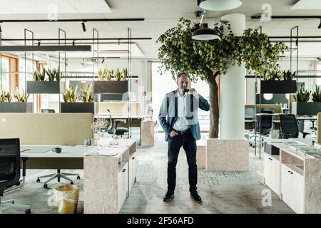 Businessman having tea while standing against tree in office Stock Photo