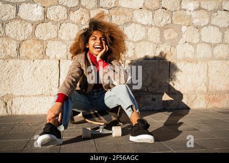 Laughing Afro woman sitting on skateboard against wall during sunny day Stock Photo