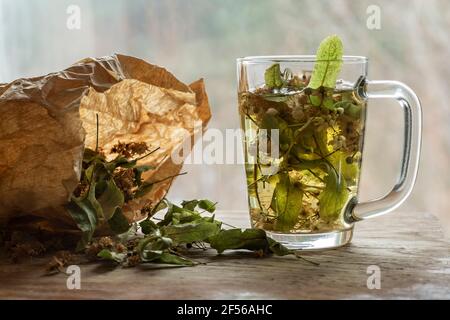 Dried linden leaves in paper bag by glass of lime tea on wooden cutting board Stock Photo