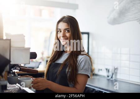 Sleepy young female starting her day on a new job as a barista. Looking happy and smiling at camera. Working in a cafe. Stock Photo