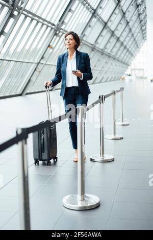 Female professional with smart phone and suitcase walking in corridor Stock Photo
