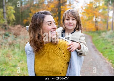Cheerful mother piggybacking smiling daughter while standing at forest Stock Photo