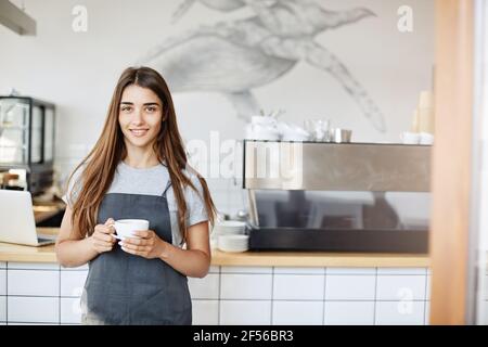 Portrait of young woman running a cafe and pastry shop standing near coffee machine looking at camera holding a cup of cappucino. Stock Photo