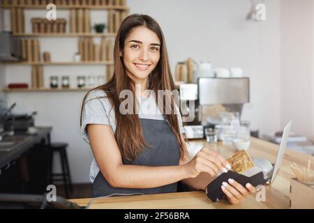 Female coffee shop employee using a credit card reader to bill the customer looking happy smiling at camera. Stock Photo