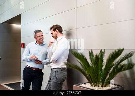 Smiling engineers using digital tablet while leaning on wall in factory Stock Photo
