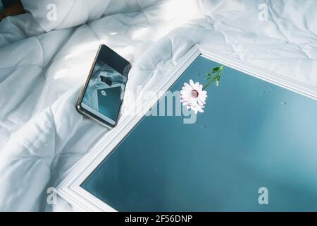 Smart phone, single daisy and mirror lying on white duvet Stock Photo