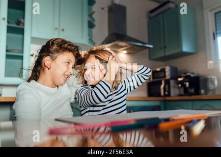 Cheerful girl playing with each other hair while sitting at dining table in kitchen Stock Photo