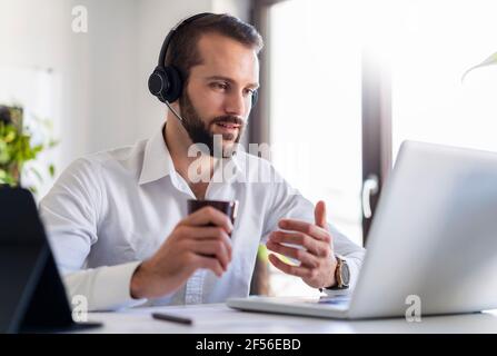 Businessman with headphones and coffee cup talking on video call over laptop at office Stock Photo