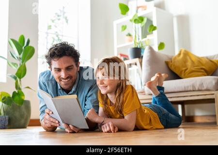 Smiling father and daughter reading book together while lying down on floor at home Stock Photo