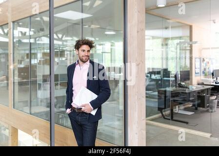 Thoughtful young male entrepreneur holding document while leaning on glass wall at office Stock Photo