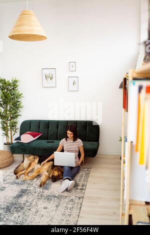 Woman with laptop stroking dog sleeping on carpet in living room