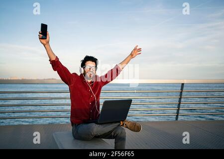 Carefree businessman with laptop raising arms while listening music through headphones on bench by the sea Stock Photo