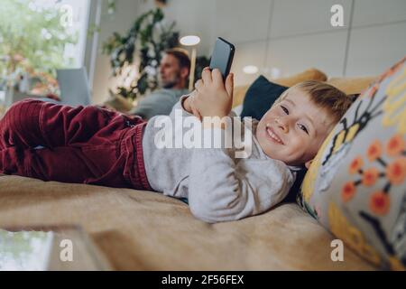 Smiling boy using mobile phone while lying on sofa at home Stock Photo