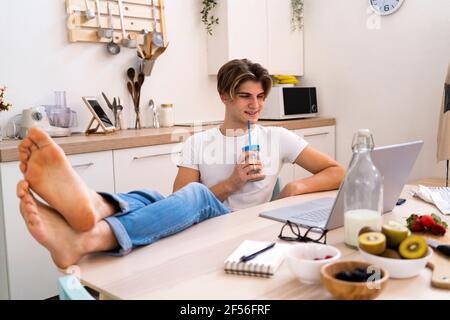 Relaxed young man drinking smoothie while sitting with laptop at table in kitchen Stock Photo