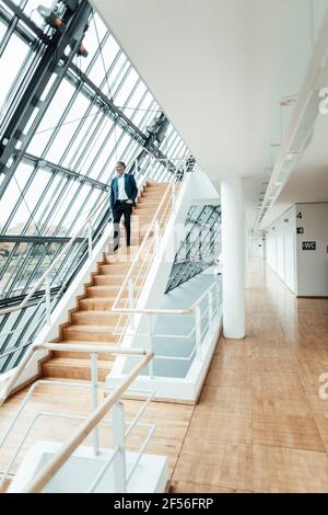 Businessman looking through window while moving down from staircase in office corridor Stock Photo