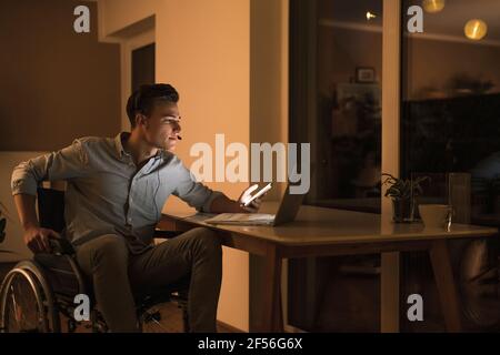 Disabled male business professional with laptop wearing headphones while sitting on wheelchair in living room Stock Photo