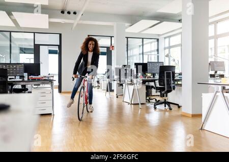 Cheerful businesswoman riding bicycle in open plan office Stock Photo