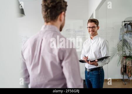 Smiling businessman with clipboard looking at male colleague in office Stock Photo