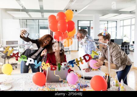 Cheerful businesswoman taking selfie with colleagues while during birthday celebration in office Stock Photo