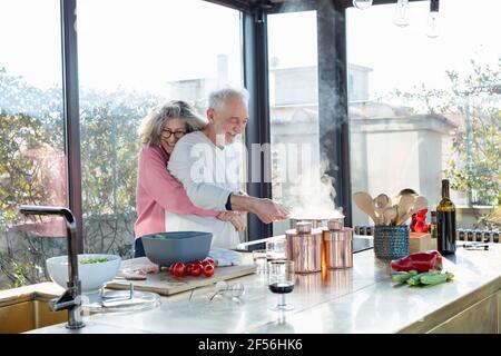 Smiling senior woman embracing man from behind in kitchen at home Stock Photo