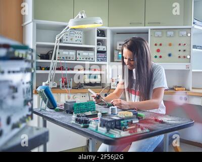 Smiling female electrician using soldering iron on circuit at electrical workshop Stock Photo
