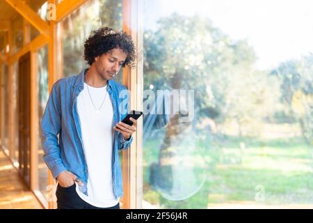 Young man using smart phone while standing at front yard window Stock Photo
